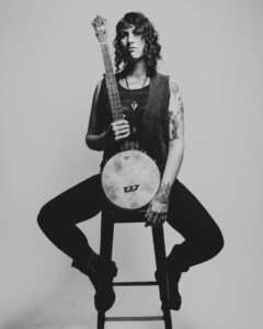 B&W photograph of Francesca sitting on a stool holding their banjo, looking at the camera.