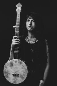 Black and white headshot of Francesca holding their banjo and looking at the camera. Their head is tilted slightly to the left.