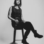 B&W photo of Francesca Mirai sitting on a stool, facing sideways with their legs crossed, holding their banjo in front of a grey backdrop. They are dressed in dark clothing, wearing jeans, a shirt with a scoop neck, a vest, a bolo tie and combat boots.