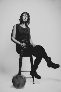 B&W photo of Francesca Mirai sitting on a stool, facing sideways with their legs crossed, holding their banjo in front of a grey backdrop. They are dressed in dark clothing, wearing jeans, a shirt with a scoop neck, a vest, a bolo tie and combat boots.